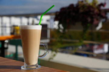 Glass cup with latte coffee on the table. Drinking tube. Cocoa in  mug on a table on the terrace. Cappuccino with milk froth. Drink at the cafe. Cocktail. Cup with coffee on the background of the city