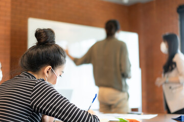 Asian businesswoman in long sleeve striped shirt wearing protective face mask sit at desk and taking notes during her asian colleagues in face masks discussing about the business plan on whiteboard.