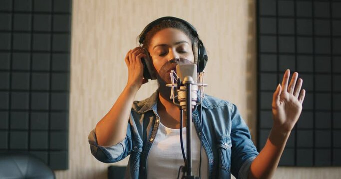 Professional vocal. Young black lady in headphones singing to microphone at sound record studio