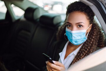 Fototapeta na wymiar Young woman in protective medical mask in the taxi car on a backseat looking out of window checking her cell phone. Health protection, safety and pandemic concept. Covid - 19.