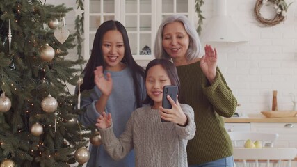 Cheerful Asian girl, mother and grandmother waving, chatting and showing Christmas tree while video calling on smartphone at home - Powered by Adobe
