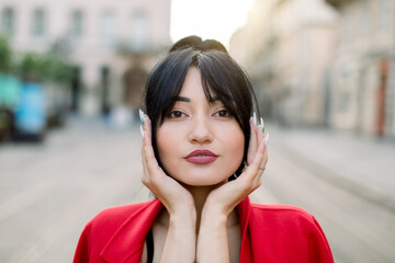 Close-up face outdoor portrait of pretty brunette Asian female model with black hair and fashion make up, leaning face on her palms, posing at camera at old city street background