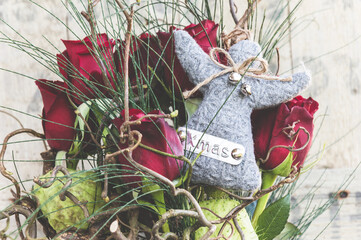 Wintry bouquet of roses in a glass vase with a gray woolen angel with XMAS lettering, old used wood as background