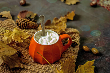 Fototapeta na wymiar Hot autumn drink in an orange cup with marshmallows, next to a knitted scarf, autumn leaves, acorns on a dark table. Autumn cozy composition