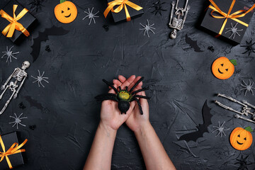 Creepy fake spider in women's palms surrounded by Halloween decorations and gifts on black background.