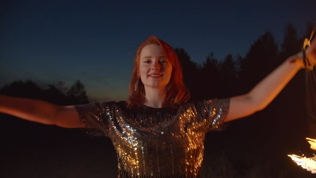 Lifestyle portrait of professional beautiful redhead female fireshow artist juggling gracefully with two fire fans in countryside at night, performing impressive fire show, expressing positive vibes.