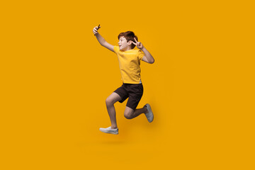 Fototapeta na wymiar Ginger caucasian boy jumping on a yellow studio wall and making a selfie using a phone gesturing the piece sign and smile