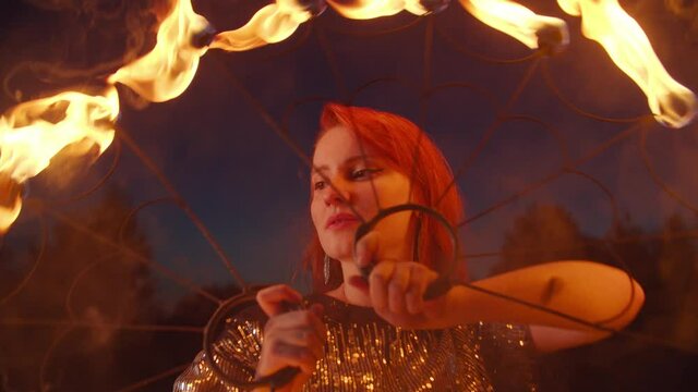 Close-up portrait of lovely redhead female fireshow performer juggling with burning fans over dark natural background, expressing confidence and magic of hypnotizing flame during fire show.