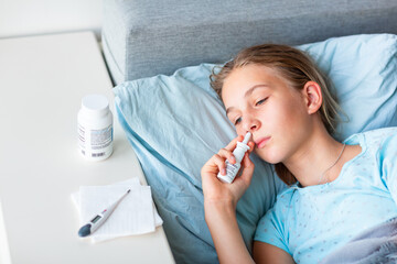 Sick teenage little girl with high fever and headache laying in bed spraying medicine into her nose