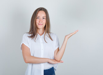 Young woman in white t-shirt, jeans welcoming or showing something and looking glad , front view.
