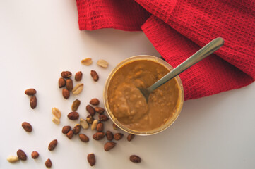 A jar of peanut butter with a spoon and randomly scattered peanuts. Ideal food for vegan.