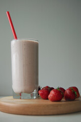 Strawberry smoothie in glass with bright red straw. One glass of smoothies and fresh strawberries are laid out randomly on wooden board on a white background.