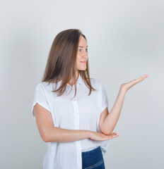 Young woman looking away with hand sign in white t-shirt, jeans and looking glad. front view.