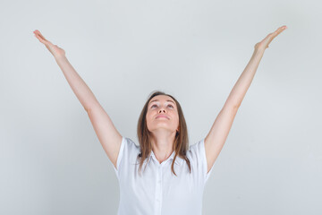 Young woman looking up with raised hands in white t-shirt and looking cheerful , front view.