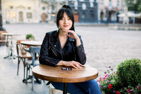 Happy young mixed raced woman sitting at cafe at the table and looking at camera. Chinese lady in black leather jacket spending free time at outdoor cafe. Old ancient buildings on the background