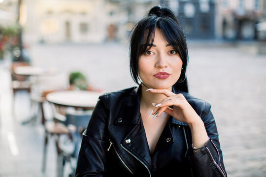 Close up portrait of elegant Asian young businesswoman in black leather jacket, sitting at the table in outdoor city cafe in old European city. Horizontal shot with cope space for text