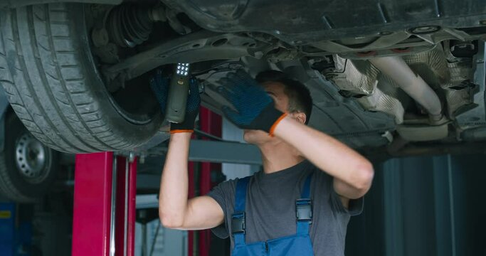 Young male mechanic repairing car suspension, standing under vehicle at garage