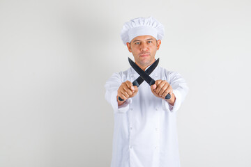 Male chef cook holding knives and looking at camera in hat and uniform front view.