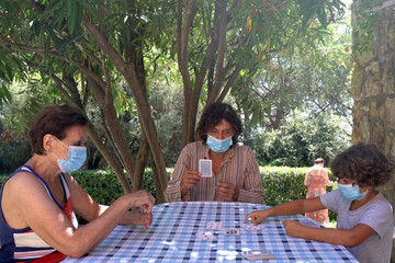 Europe, Italy , September 2020 - Grandmother , father and children boy  five years at home during quarantine due Covid-19 Coronavirus  - lifestyle in apartment - playing card with mask in the garden