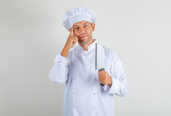 Male chef cook holding knife for meat and thinking in uniform and hat and looking positive. front view.