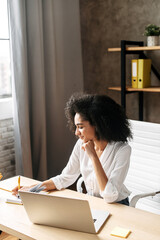 A young african woman looks at laptop screen and takes a note sitting in contemporary office....