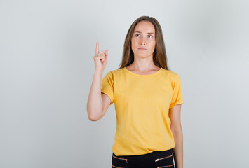Young woman in yellow t-shirt, pants pointing finger up and looking pensive , front view.