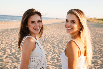 Two young caucasian women smiling at camera while walking on beach