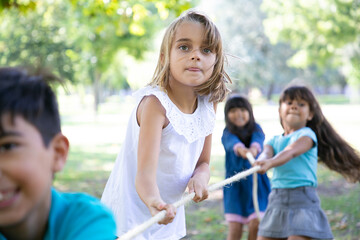 Excited girl enjoying outdoor activities with classmates, playing tug-of-war with friends. Group of children having fun in park. Childhood concept