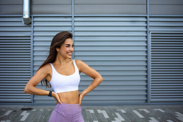 Fototapeta na wymiar Female fitness model in white top and purple leggings is posing outdoors. Full length portrait of a young fitness woman in sportswear posing and jumping