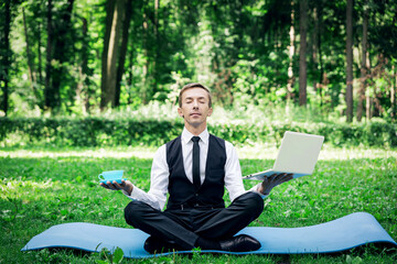 Attractive young male workaholic in a business suit and tie, sits in a lotus position in the park,...