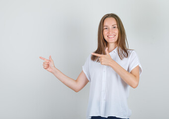 Young woman pointing fingers to side in white t-shirt and looking cheerful. front view.