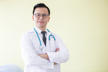 portrait asian doctor wearing eyeglass in suit uniform arm cross posing and looking at camera yellow background in hospital.