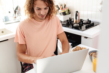 Caucasian handsome pleased guy using laptop while having breakfast