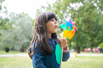 Adorable black haired girl playing in park, holding pinwheel and blowing on toy. Medium shot, side view. Children outdoor activity concept