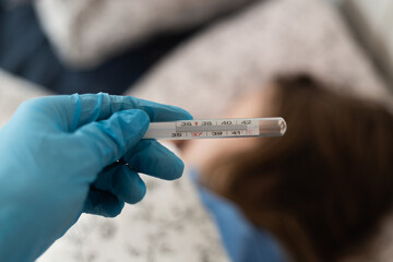 Close up of medical thermometer with blue rubber glove against sick woman lying in bed. 