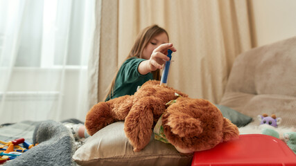 A cute small girl making and injection to a teddybear playing doctor sitting on a sofa in a bright cosy room