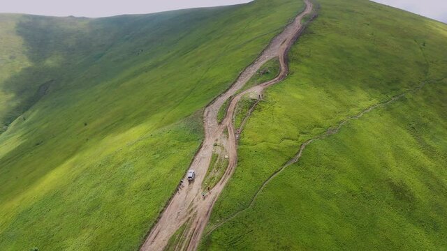 Aerial drone view of a winding dirt road crossing a lush green mountain ridge, with rolling hills, dramatic natural landscape, and soft daylight. Scenic highland terrain captured from above