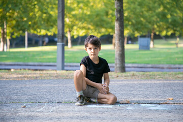Serious cute boy sitting and drawing on asphalt with colorful pieces of chalks, looking at camera. Front view. Childhood and creativity concept
