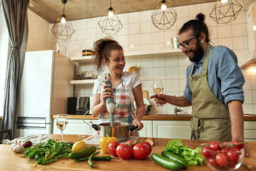 Italian man, chef cook holding a glass of wine while woman using hand blender. Cheerful couple...