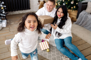 Obraz premium Mixed east asian family parents and their little daughter hugging and giving each other gifts by the christmas tree