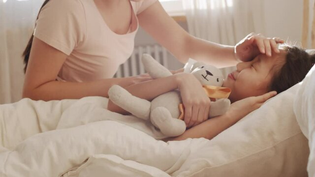Beautiful Asian mother waking up cute little daughter while sleeping with bear toy in bed, smiling and talking with her in the morning at home
