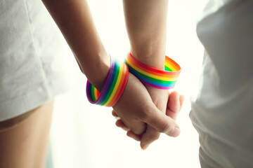 Closeup lesbian couple holding hands with a rainbow-patterned wristban on their wrists.Concept of LGBT, activism, community and freedom. - obrazy, fototapety, plakaty