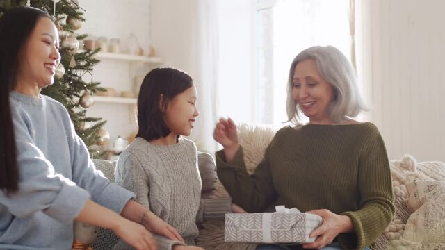 Beautiful senior woman getting Christmas present from cheerful Asian daughter and little granddaughter and then untying ribbon on box while sitting together on couch in cozy living room