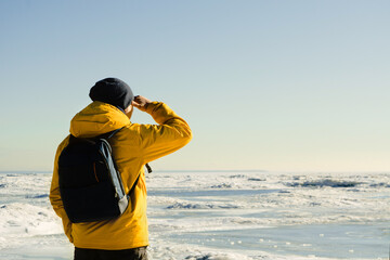 Obraz premium One faceless man in yellow raincoat with backpack standing on frozen sea and looking far on nature. Copy space. Tourist hiking. Freedom of travel concept. Unrecognizable male