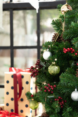 Selective focus of artificial christmas tree decorated with siver & golden ball, pine cone, and red cherry over blurry background of poka dot pattern present box and windows