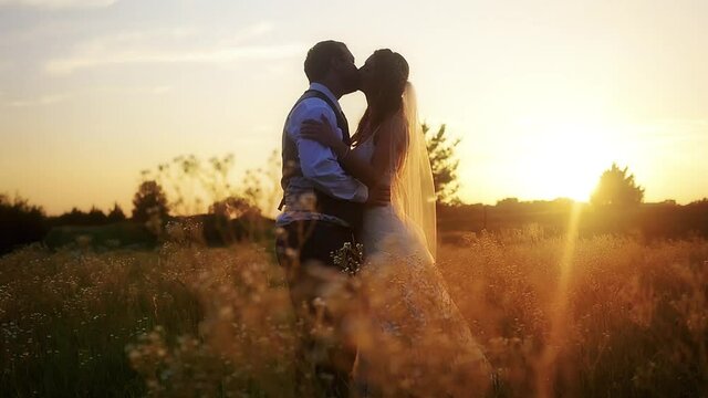 A bride and groom kiss in a Kansas meadow full of little wildflowers as the warm sun sets behind them on the horizon