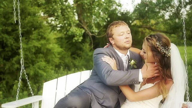 A groom goofing around trying to gross out his bride on a white bench swing in a green field