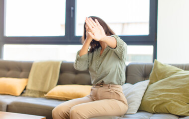 young latin woman covering face with hand and putting other hand up front to stop camera, refusing...