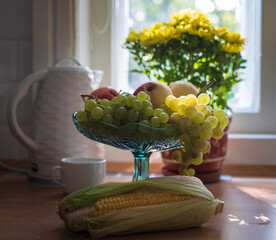 Autumn style life in the kitchen in a light rustic style, with grapes in a vase against the background of .yellow flowers of a white teapot and a window.