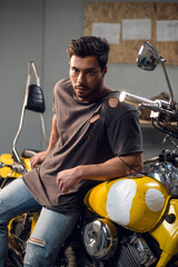 A handsome young motorcyclist in jeans and a t-shirt poses for a photo sitting on a bike in his garage.